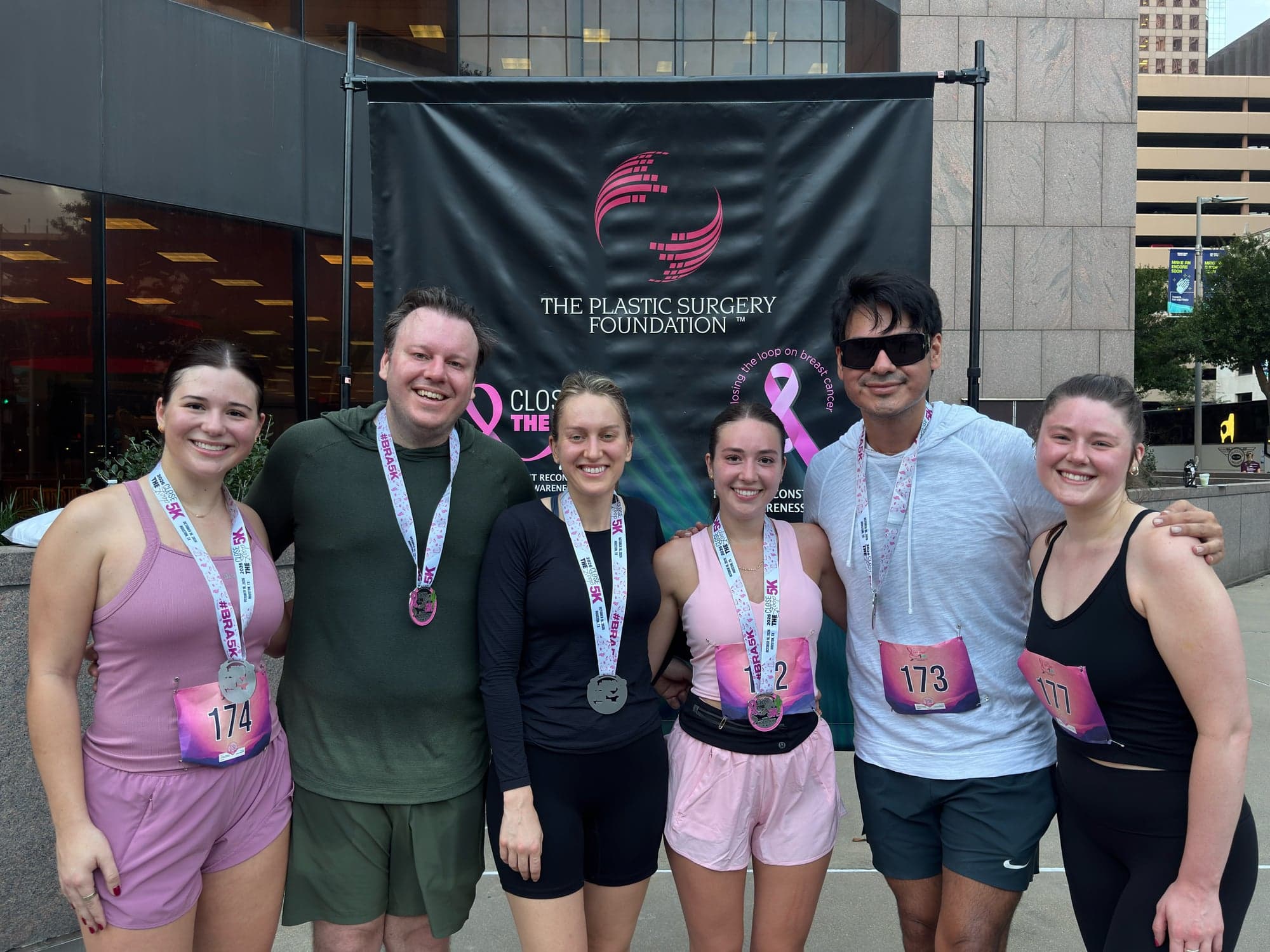 Six Vitenas Cosmetic Surgery team members wearing finisher medals in front of the Close the Loop 5K backdrop.